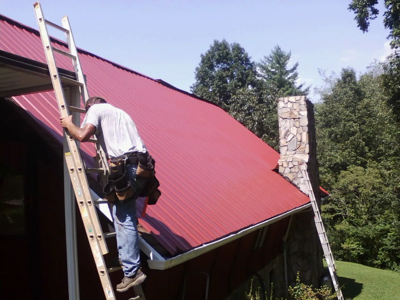 Roofer on a ladder beside a red metal roof and stone chimney — Soffit & Fascia Repair by Roofing Brothers in Laguna Woods